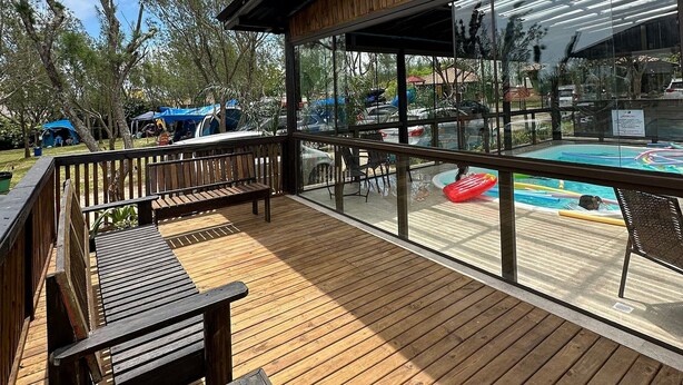 A wooden deck with a pool area, including a bench and chairs, and a view of the pool through large windows. - Pousada Cardoso Surf Camping - Laguna - SC (Laguna)