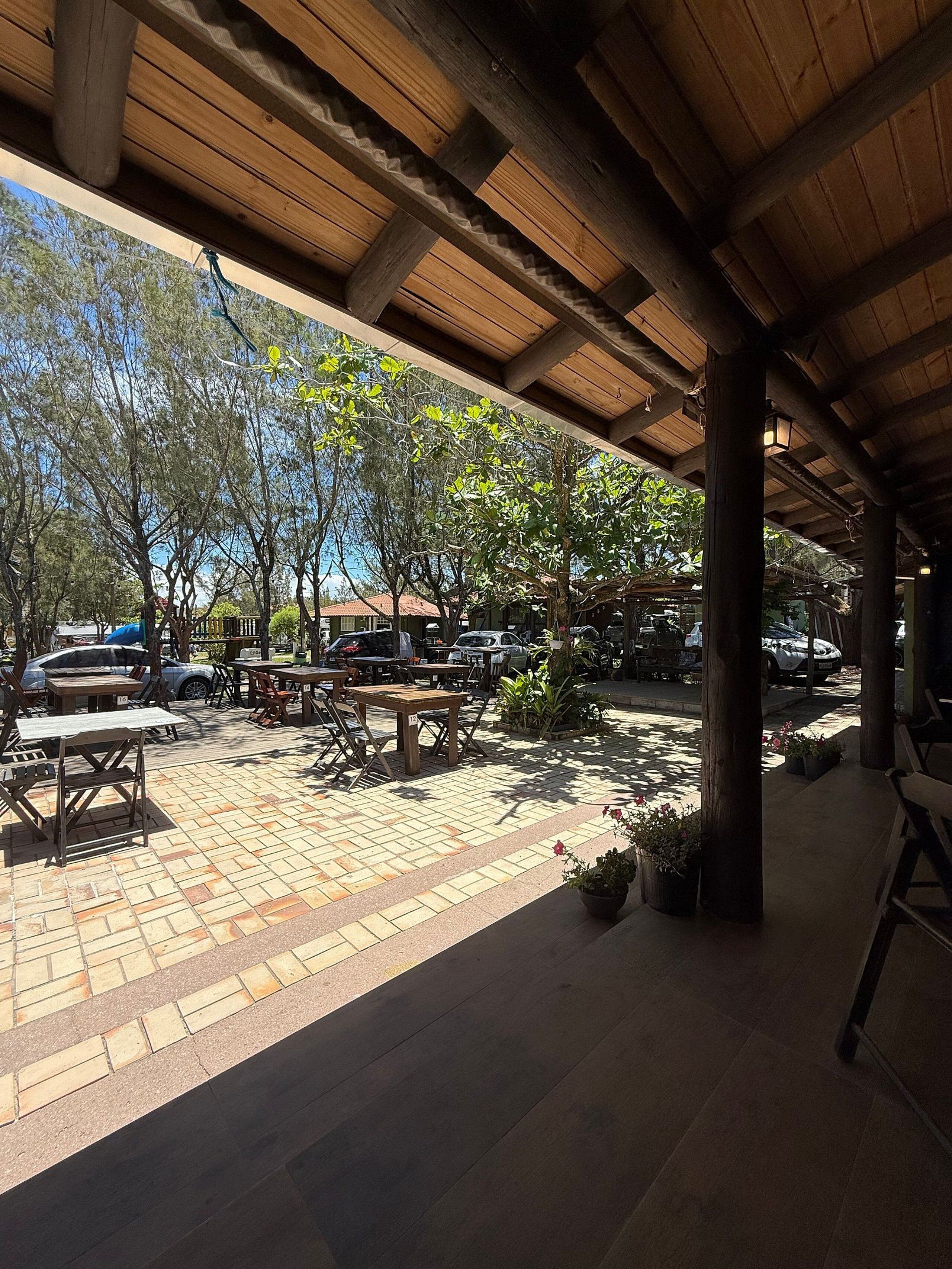 An outdoor patio area with wooden tables and chairs, surrounded by potted plants and trees.