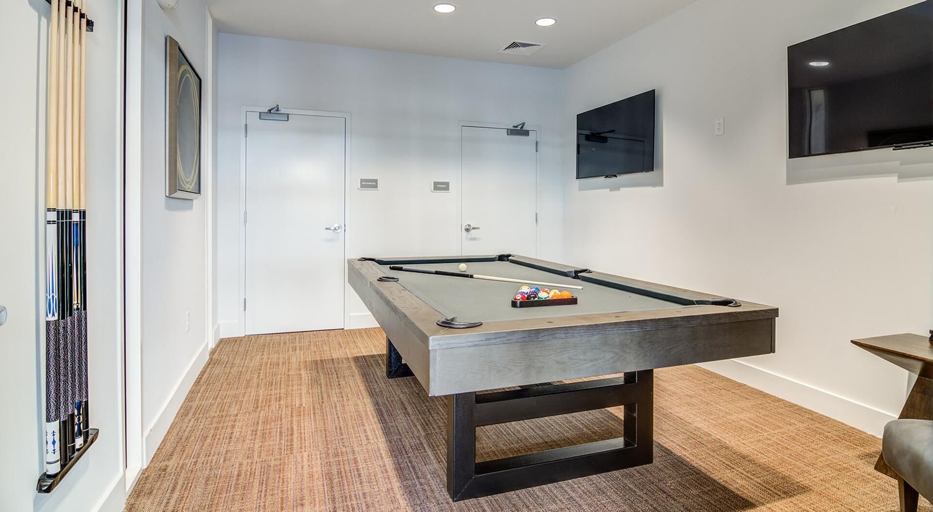 Une table de billard dans une pièce dont le sol est recouvert d’une moquette, les murs sont blancs et un téléviseur est fixé au mur.