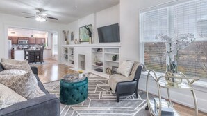A modern living room with a sofa, armchair, and ottoman, a TV, and a window with blinds. - Carson Flats Apt #6 | 1BR • Near BOK• OSU Med•Dtwn (Tulsa)