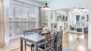 A dining area with a wooden table and chairs, a pendant light, and a view of the city through large windows with blinds. - Carson Flats Apt #6 | 1BR • Near BOK• OSU Med•Dtwn (Tulsa)