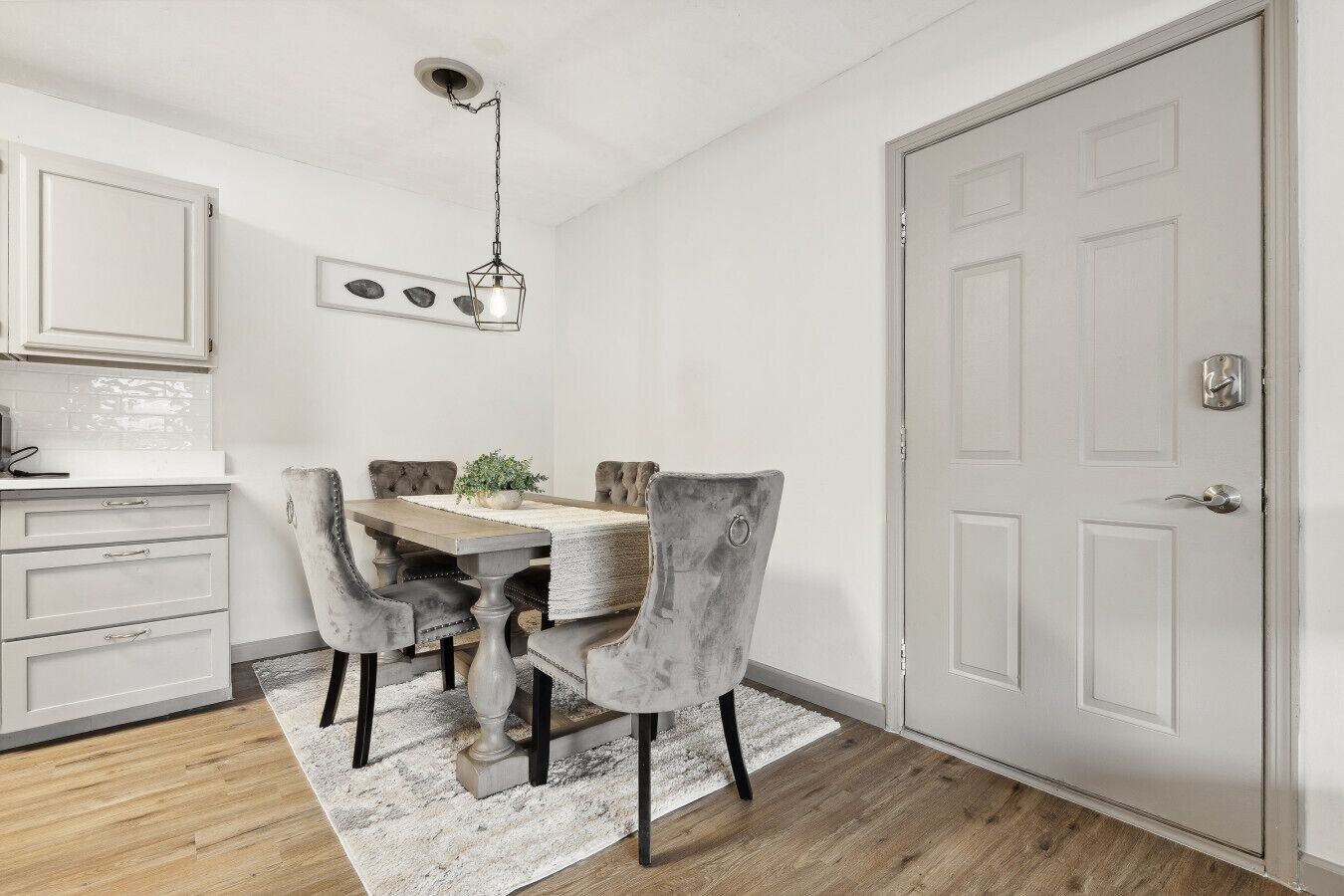 Een eetruimte met een houten tafel, grijs gestoffeerde stoelen, een wit tapijt en een witte deur met een zilveren handgreep.