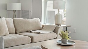 A living room with a beige sectional sofa, a side table with a lamp, and a potted plant on a tray. - Landing | Incredible 1BD, Clubhouse, Pool (Decatur)