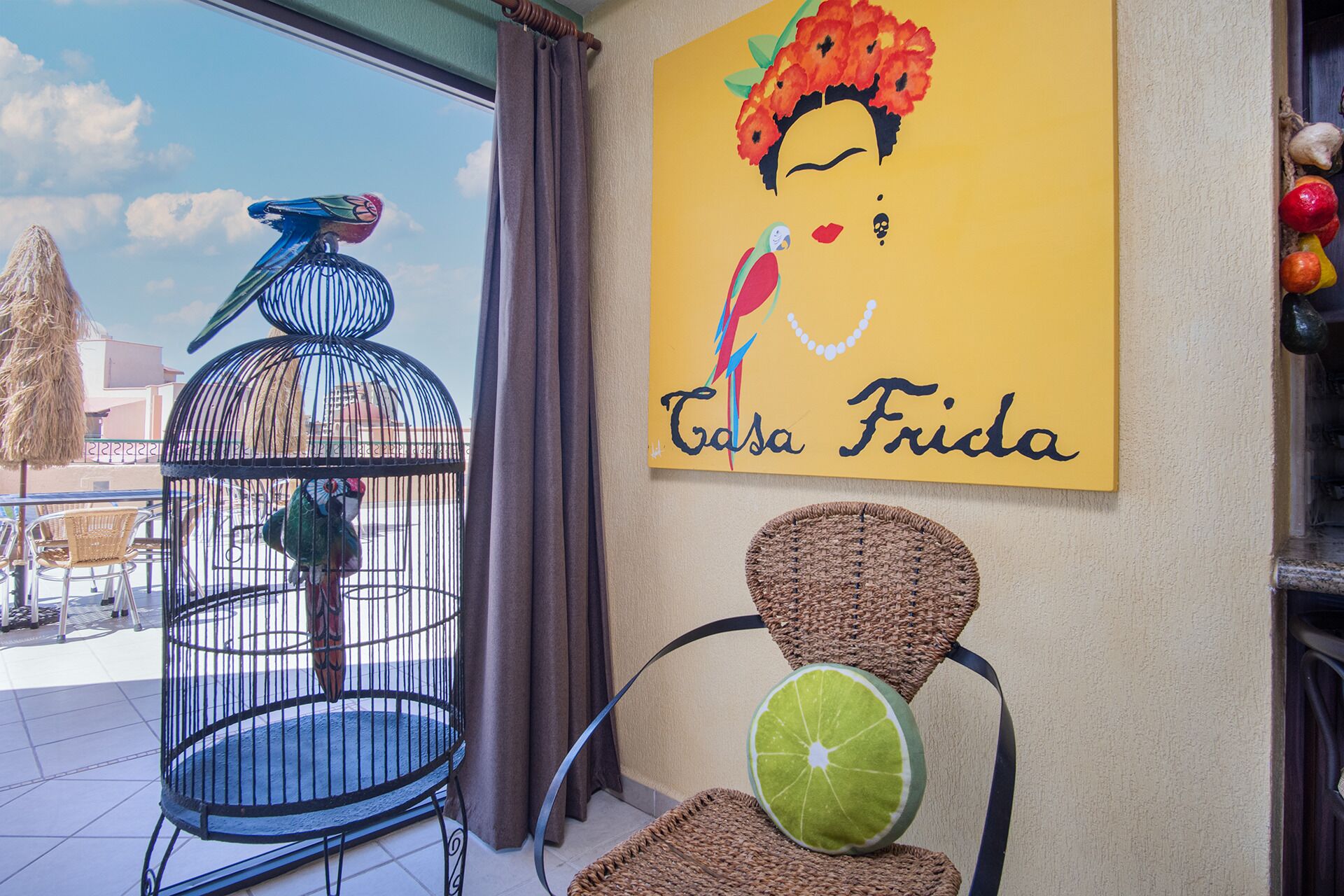 Huone, josta on näkymä rakennukseen, papukaijan häkki, seinä, jossa on kyltti, jossa lukee ”Casa Frida”, ja punotut tuolit.