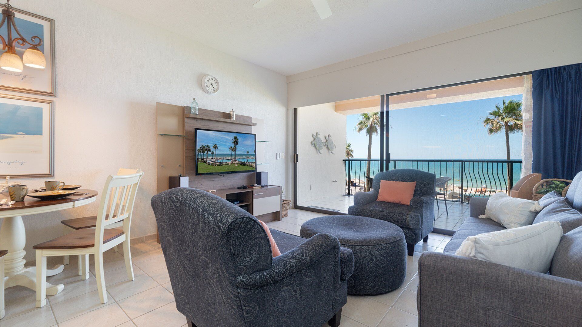 Un salón moderno con un televisor de pantalla plana, un comedor con una mesa y sillas blancas, y un balcón con vistas a la playa.