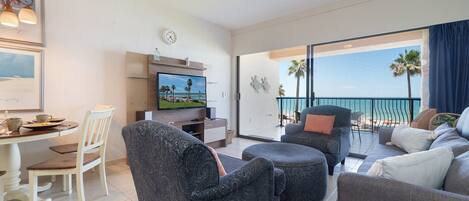 Un salón moderno con un televisor de pantalla plana, un comedor con una mesa y sillas blancas, y un balcón con vistas a la playa.