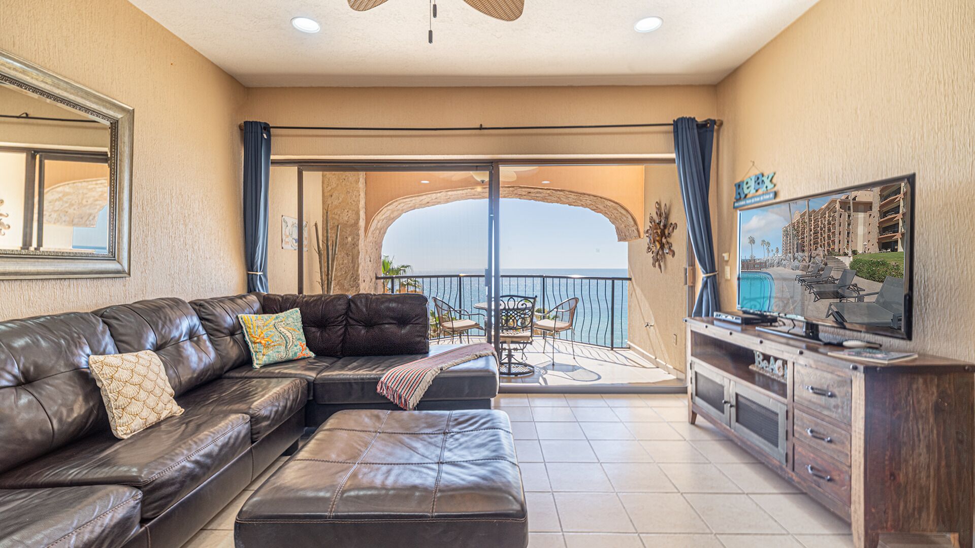 Un salon avec un canapé en cuir, un pouf et un téléviseur posé sur un meuble en bois. On y trouve également un balcon avec vue sur la mer et un bâtiment en arrière-plan.