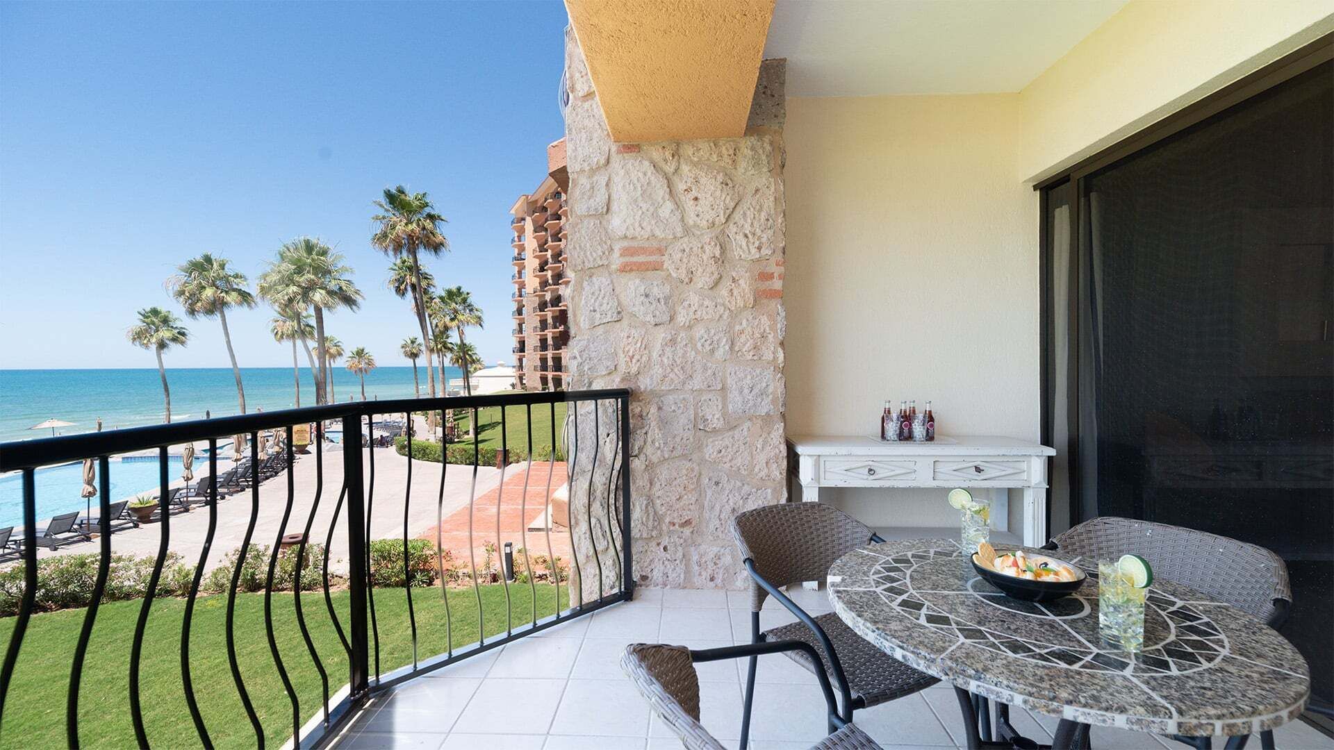 A balcony with a view of the ocean, a dining table with chairs, and a small white table with bottles.