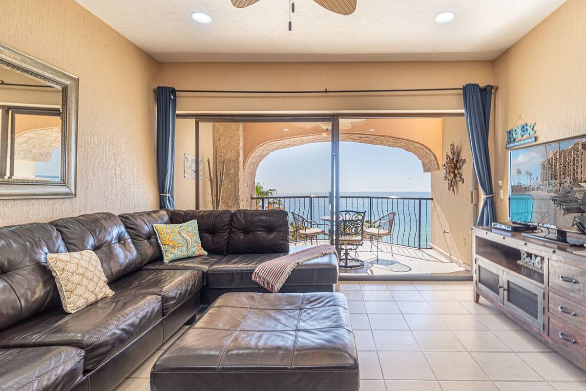 Un salon avec un canapé en cuir, un pouf et un téléviseur posé sur un meuble en bois. On y trouve également un balcon avec vue sur la mer et un bâtiment en arrière-plan.