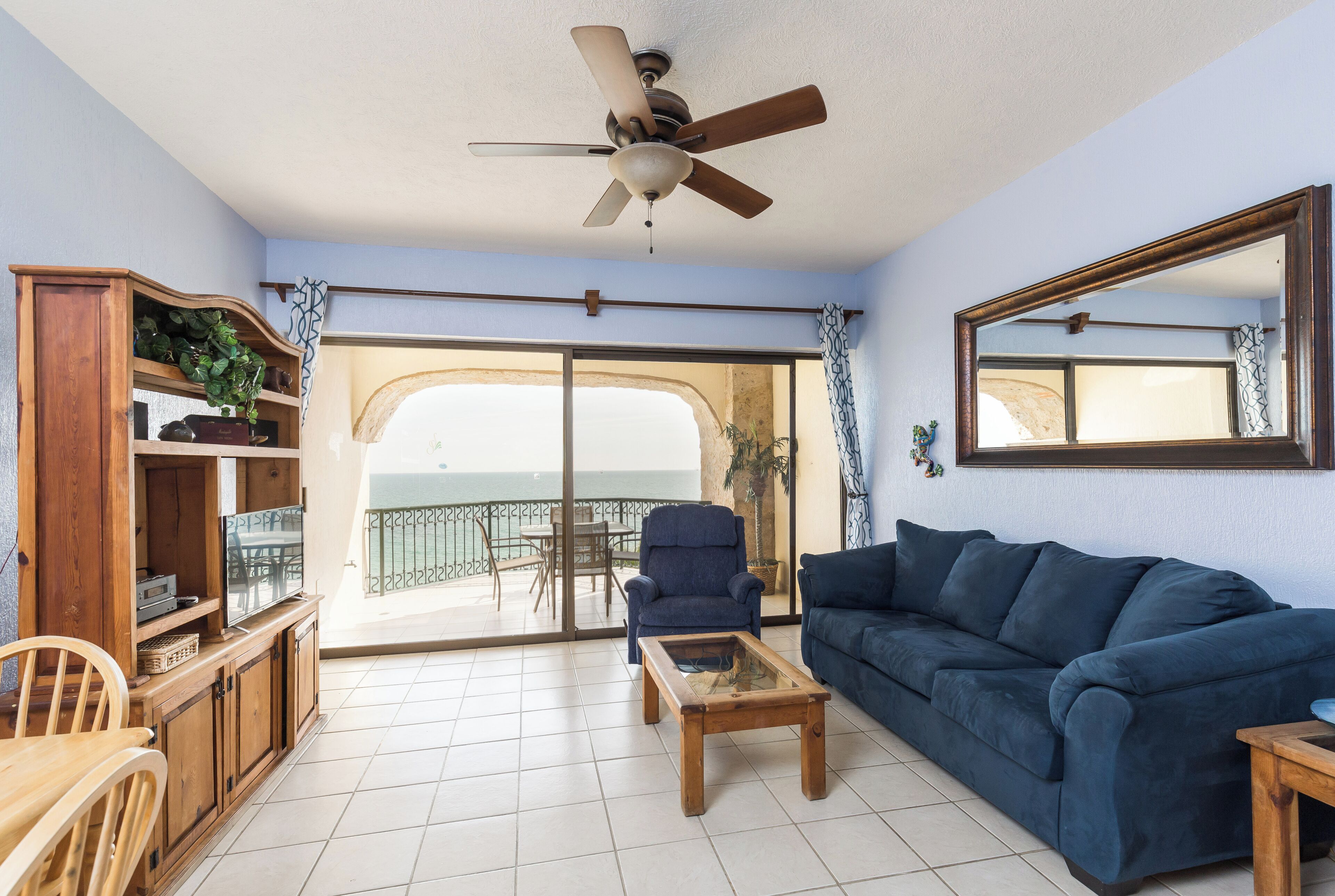 Uma sala de estar com sofá azul, mesa de centro de madeira e ventilador de teto. Há um armário de madeira com prateleiras e uma janela grande que oferece uma vista.