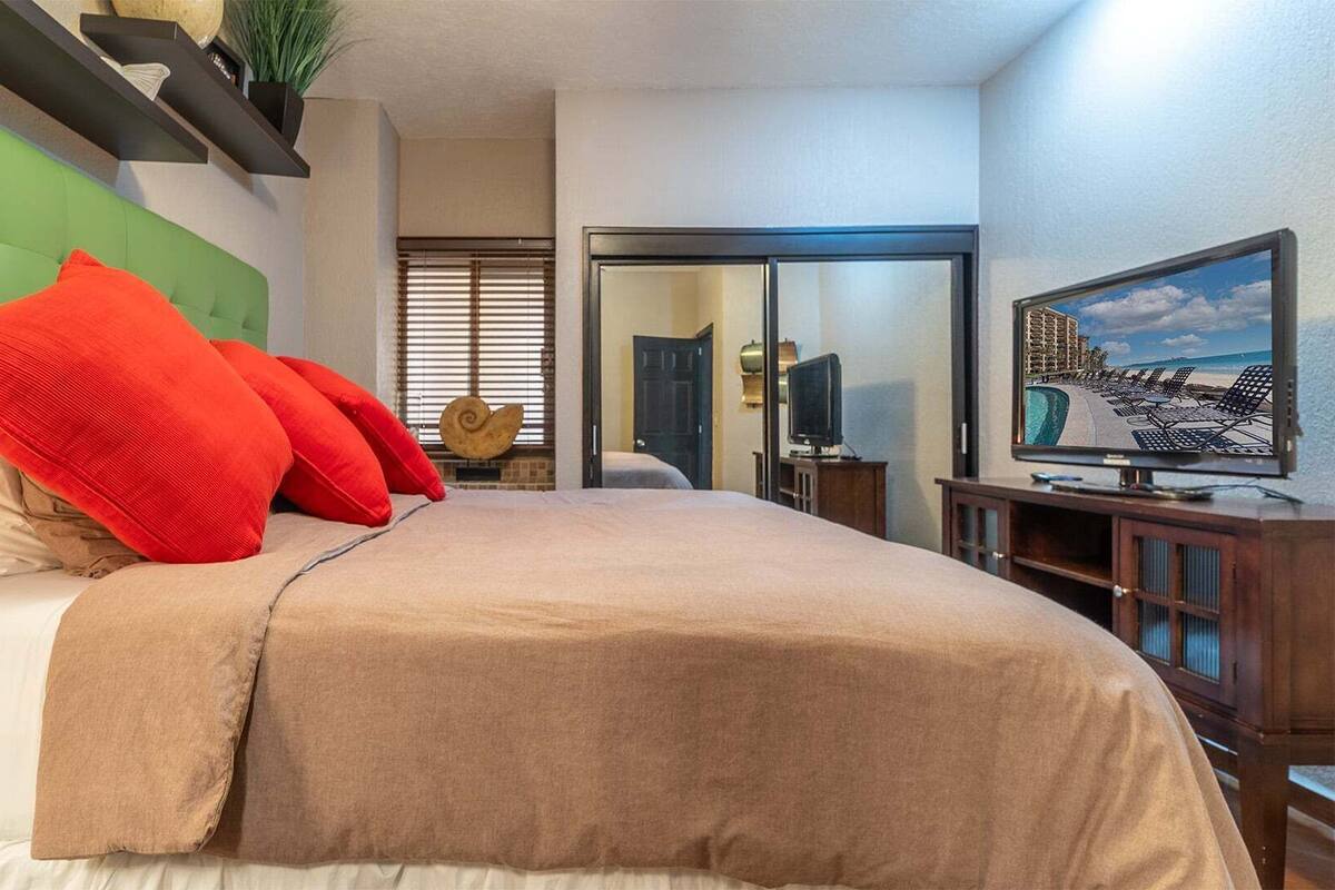 Hotelkamer met een groen bekleden hoofdeinde, een groot bed met beige en rode beddengoed, een televisie op een houten standaard en uitzicht op een zwembad en hotelgebouw door een glazen deur.