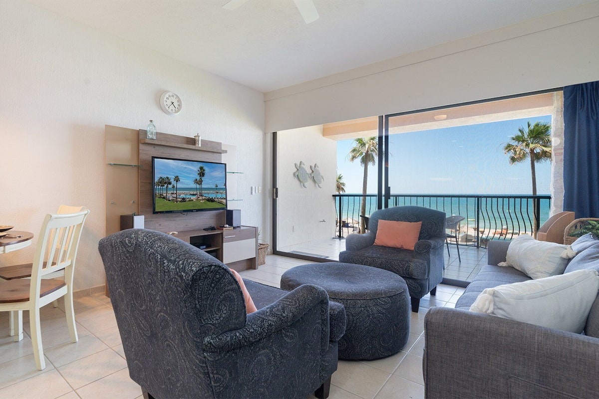 Un salon moderne doté d’un téléviseur à écran plat, d’un coin repas avec une table et des chaises blanches, et d’un balcon offrant une vue sur la plage.