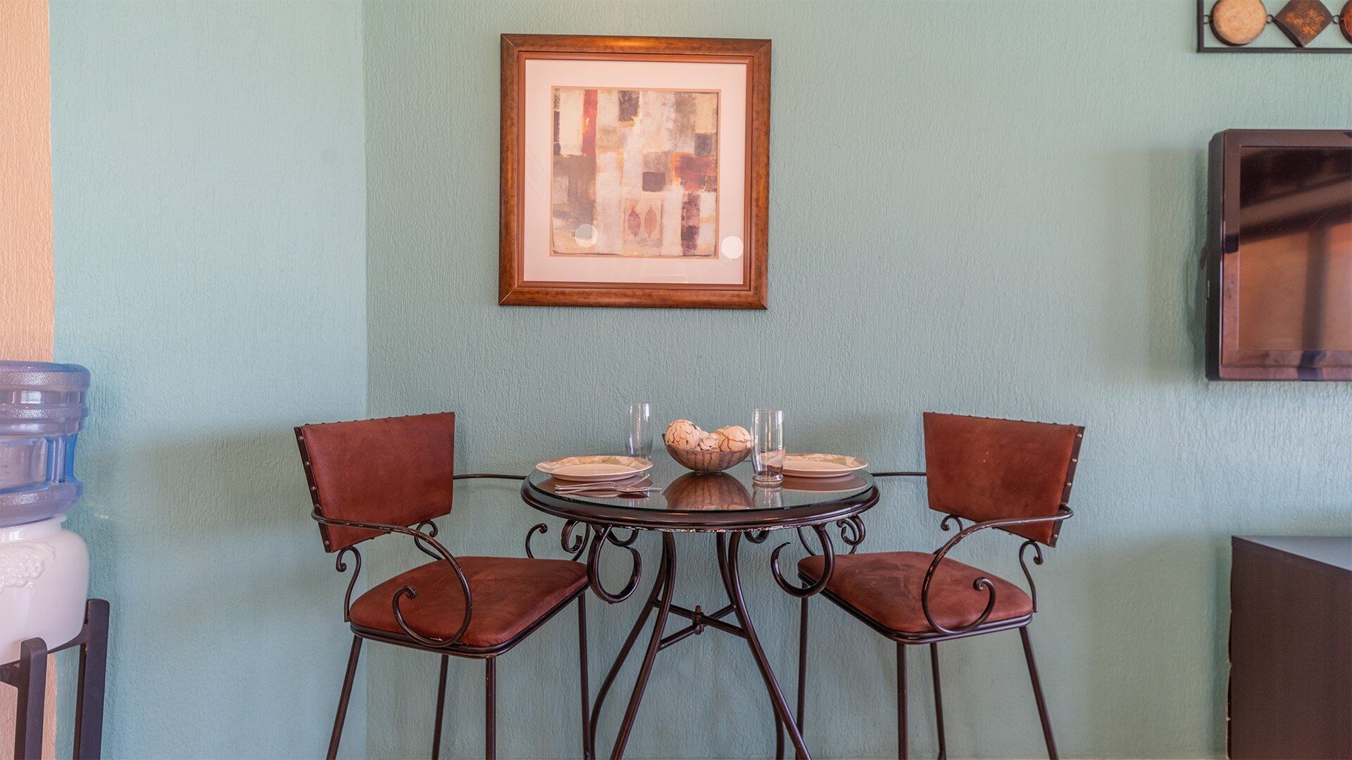 Een eetgedeelte met een ronde tafel en twee stoelen, een lijst met een abstract schilderij aan de muur, en een waterdispenser aan de linkerkant.