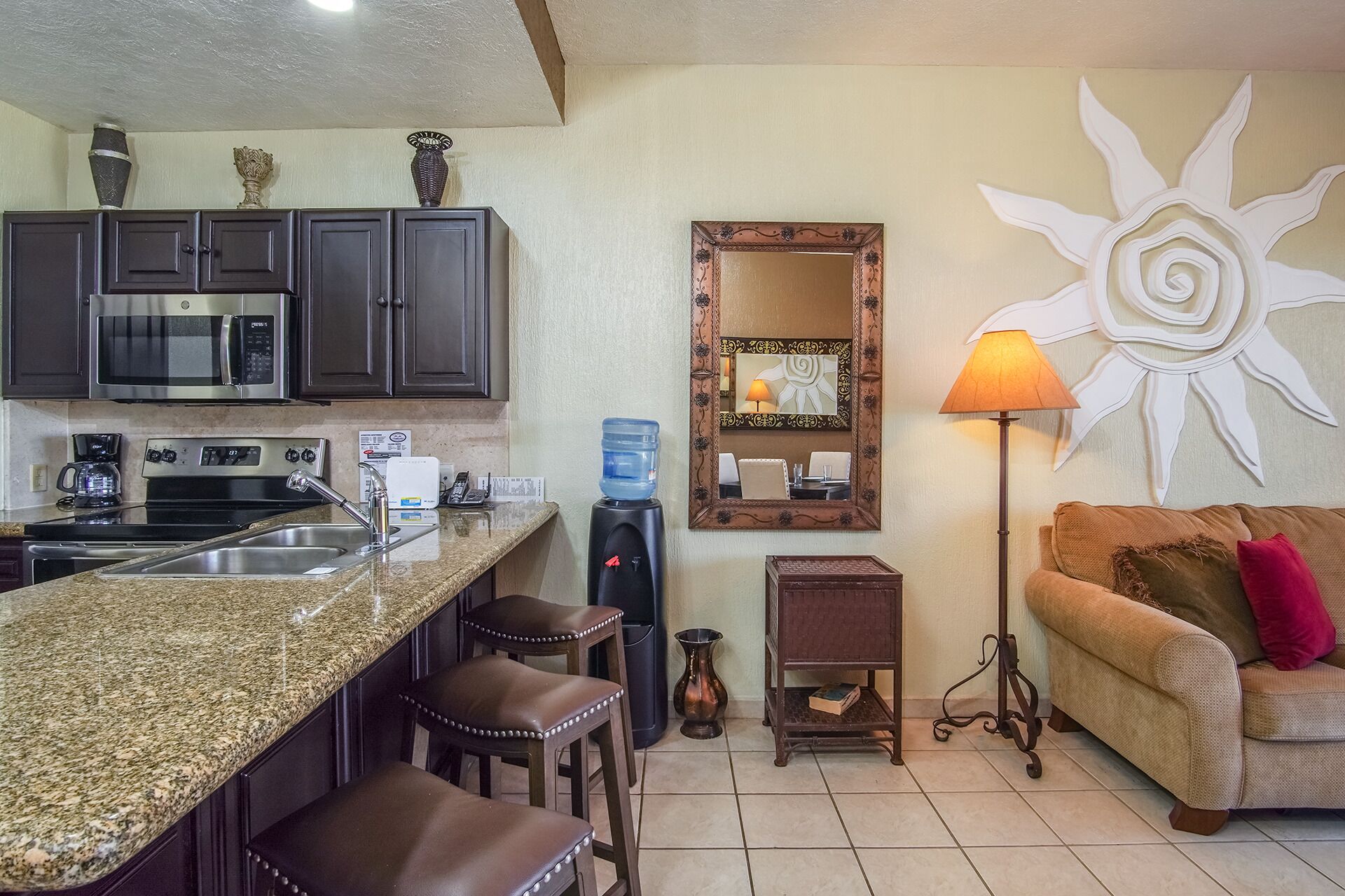 Cocina compacta con microondas, estufa y taburetes de bar. Un espejo grande y un adorno de sol están en la pared.