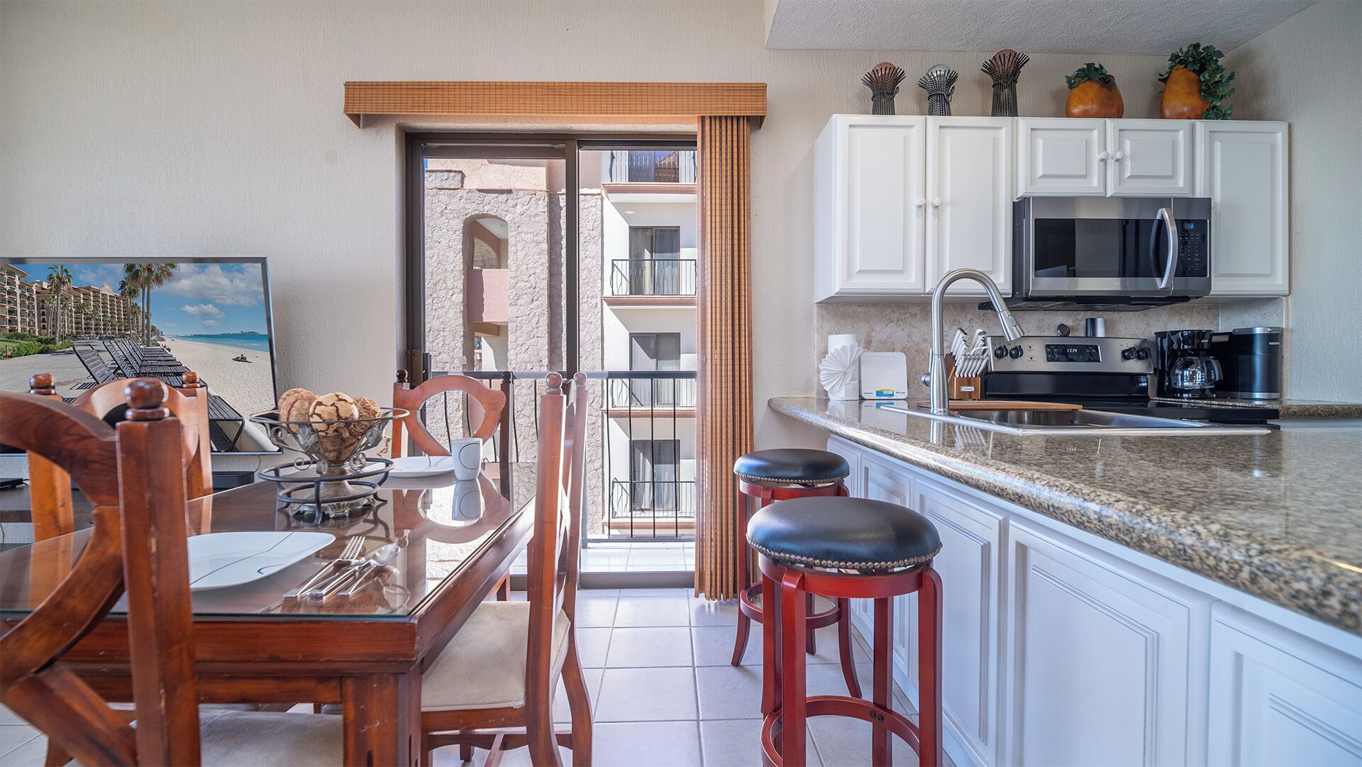 Una cocina con armarios blancos, un microondas de acero inoxidable y una cafetera. Hay una zona de comedor con una mesa de vidrio y sillas de madera.