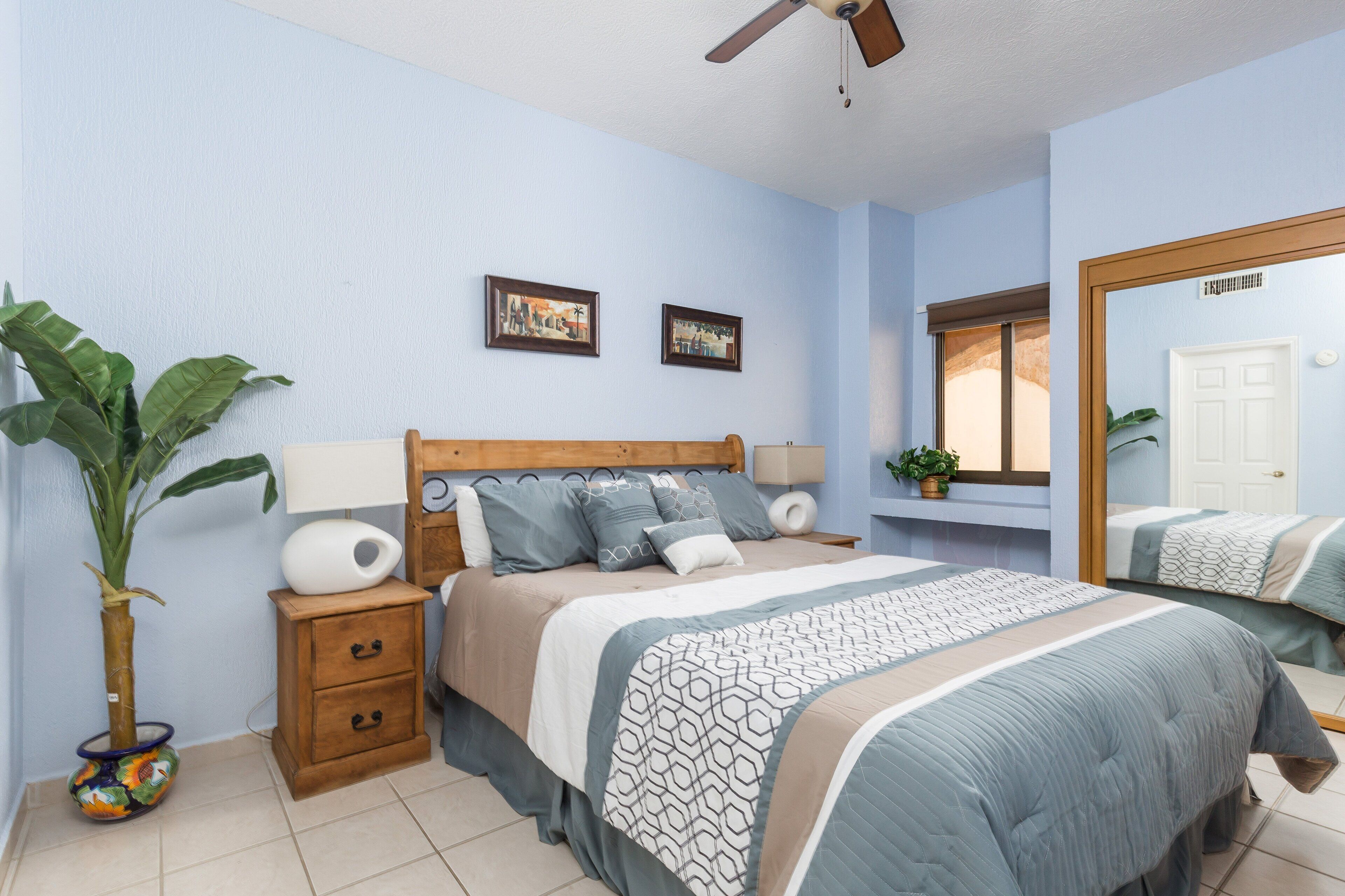 Une chambre à coucher comprenant un lit, des tables de chevet, un ventilateur de plafond, un grand miroir et une plante en pot.