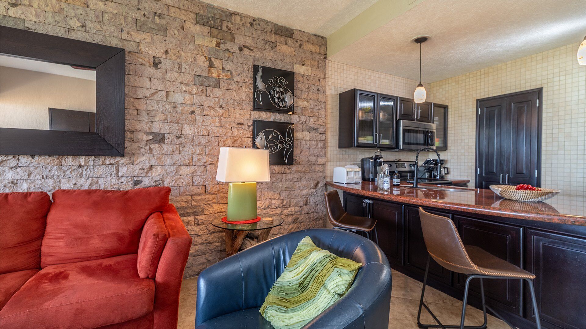 A modern kitchen with dark cabinets, a microwave, and a coffee maker. There is a red sofa and a blue chair with a green cushion. A lamp is on a small table.
