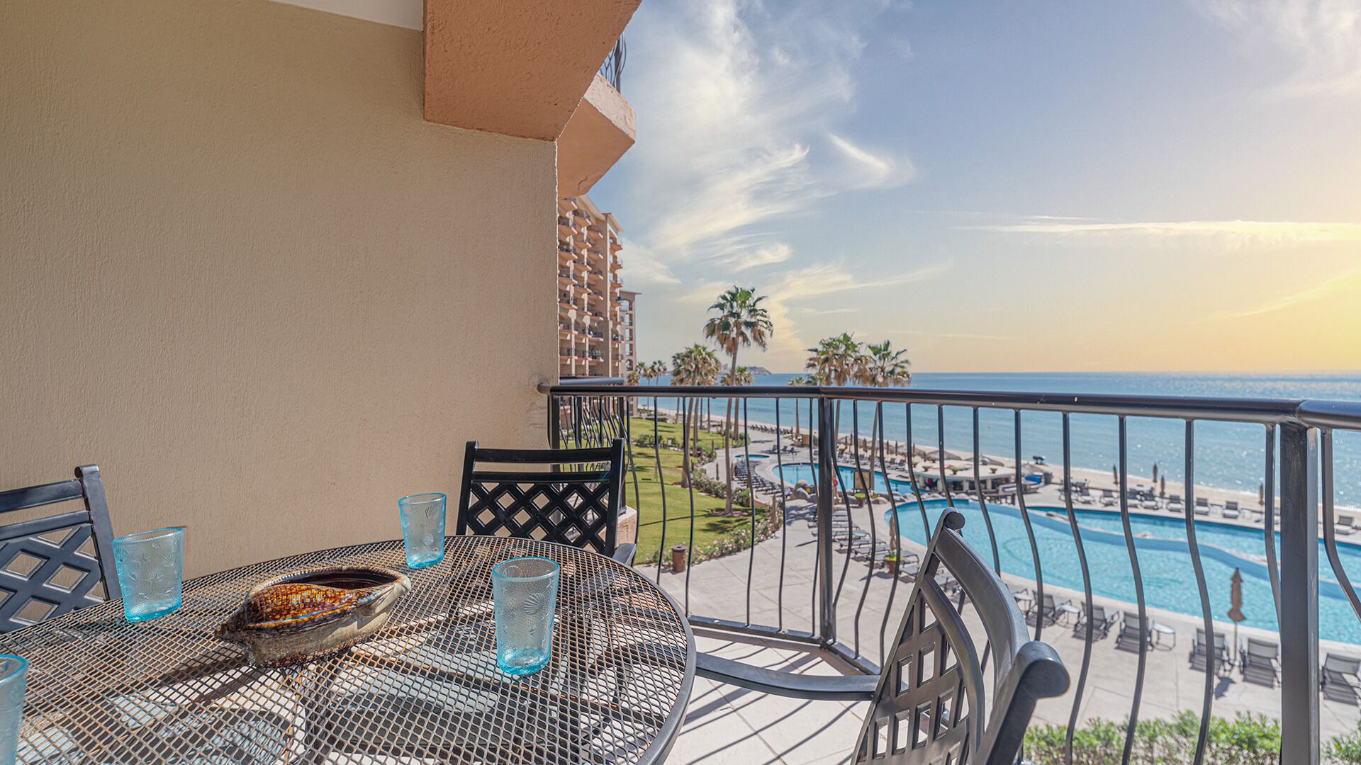 A balcony with a round table, two chairs, and a bowl of fruit. A pool and beach are visible in the background.