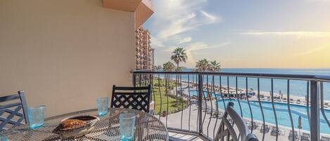 A balcony with a round table, two chairs, and a bowl of fruit. A pool and beach are visible in the background.