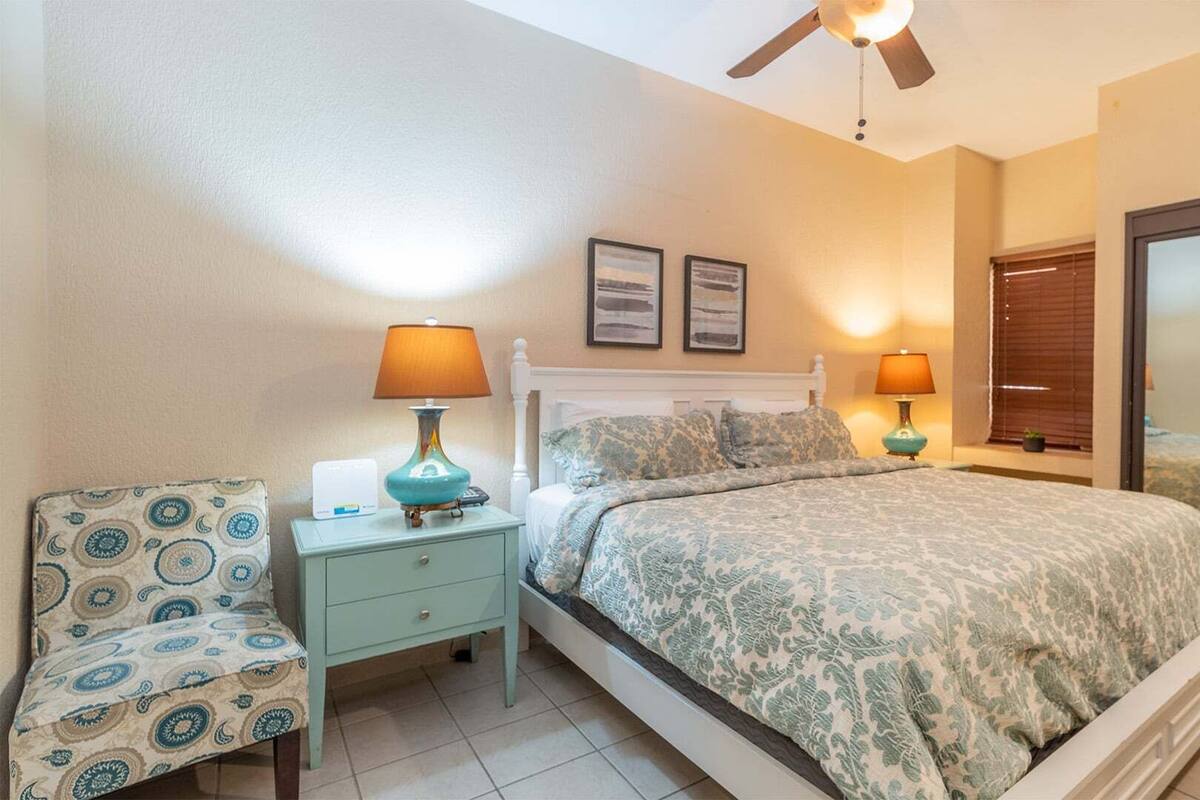 Une chambre à coucher avec un lit, deux tables de chevet avec des lampes, une chaise, un ventilateur de plafond et deux tableaux encadrés accrochés au mur.