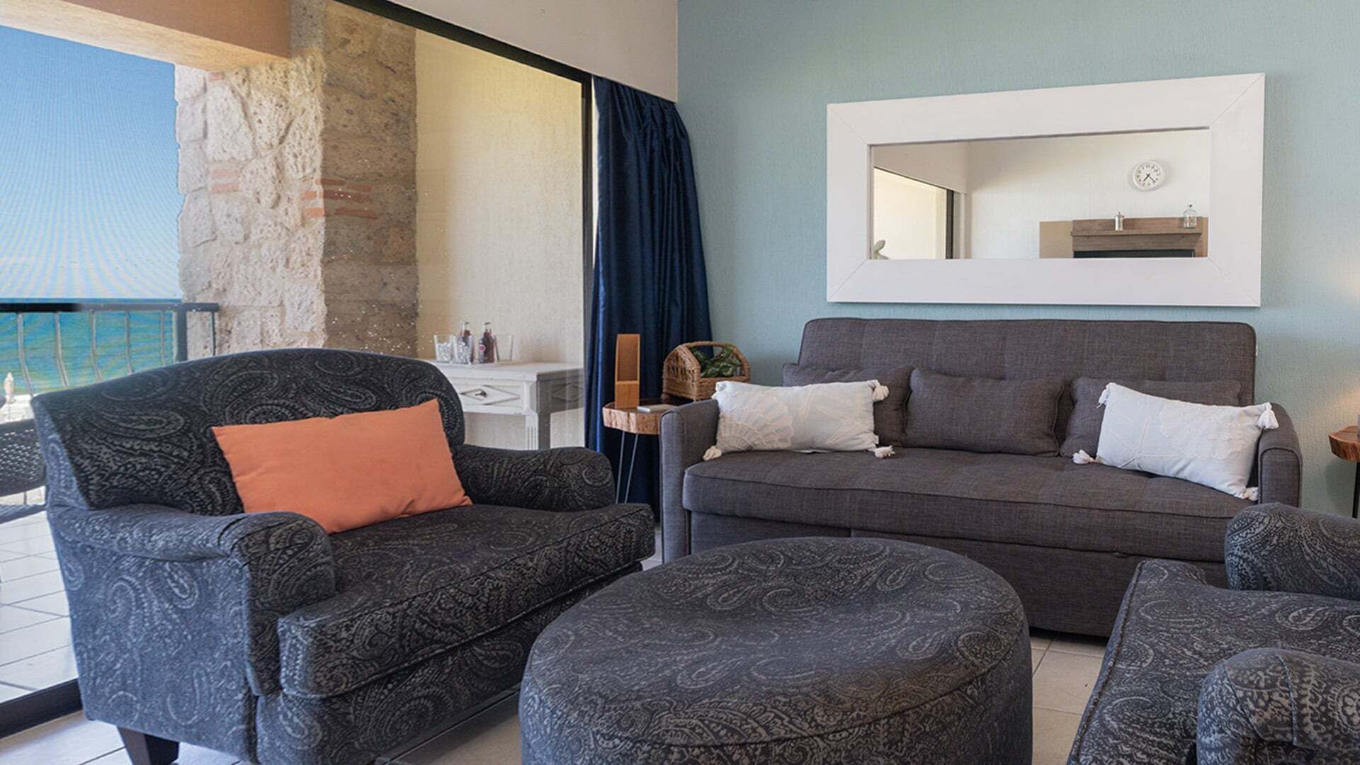 Una sala de estar moderna con un sofá gris, un reposapiés a juego y sillones estampados. Se ve una mesita auxiliar con una lámpara y objetos decorativos.