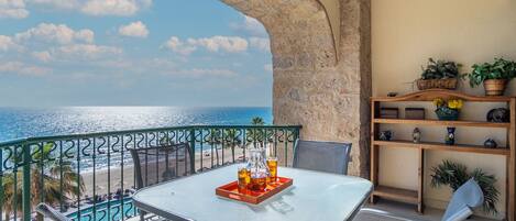 A balcony with a table and chairs, overlooking a beach and the sea.