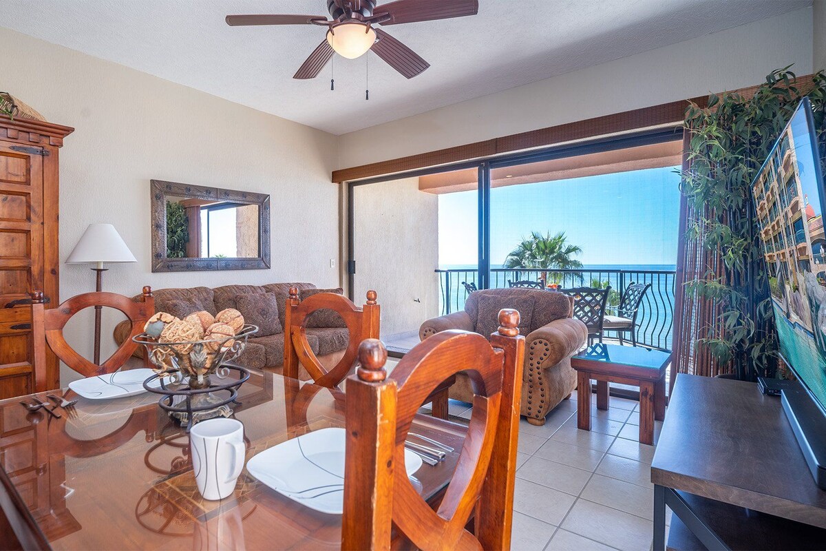 A spacious living area with a wooden cabinet, a dining table with chairs, a sofa, and a balcony with a view of the ocean.