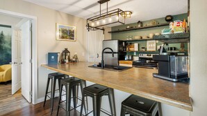Une cuisine dotée d’un plan de travail en bois, de tabourets de bar, d’un réfrigérateur, d’une machine à café et d’étagères où sont rangés de la vaisselle et des objets de décoration.