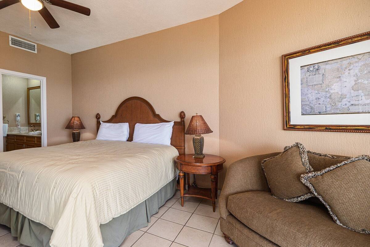Een hotelkamer met een groot bed, een houten hoofdbord, een kleine ronde tafel met een lamp, een bruine bank en een inlijst kaart aan de muur.