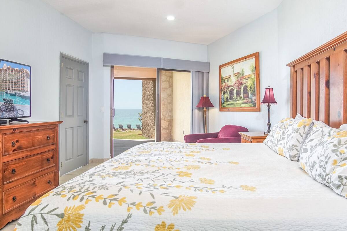 Een slaapkamer met een groot bed, een houten dressoir, een bureau, een stoel, een schilderij en uitzicht op zee.