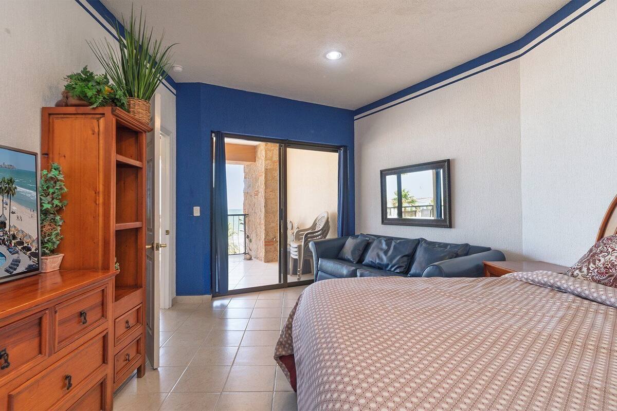 Een slaapkamer met een bed, een houten dressoir, een blauwe wand, een balkon met uitzicht op het strand en een lijst met een afbeelding.