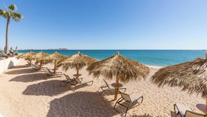 Ein Strand mit Reihen von Liegestühlen und reetgedeckten Sonnenschirmen, einer Palme und freiem Blick auf das Meer.