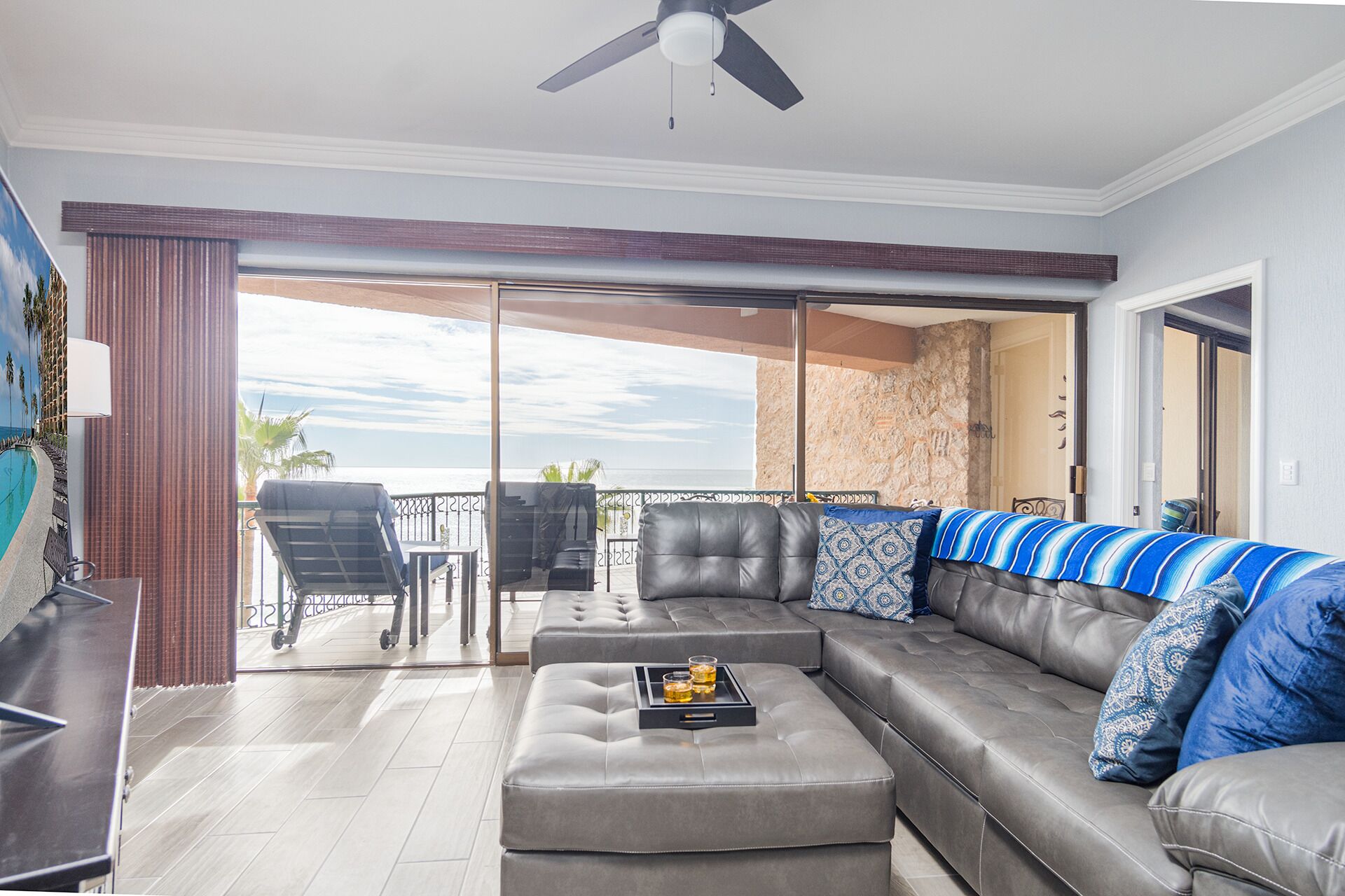 A modern living room with a grey sectional sofa, a coffee table with drinks, and a balcony with a view of the ocean.