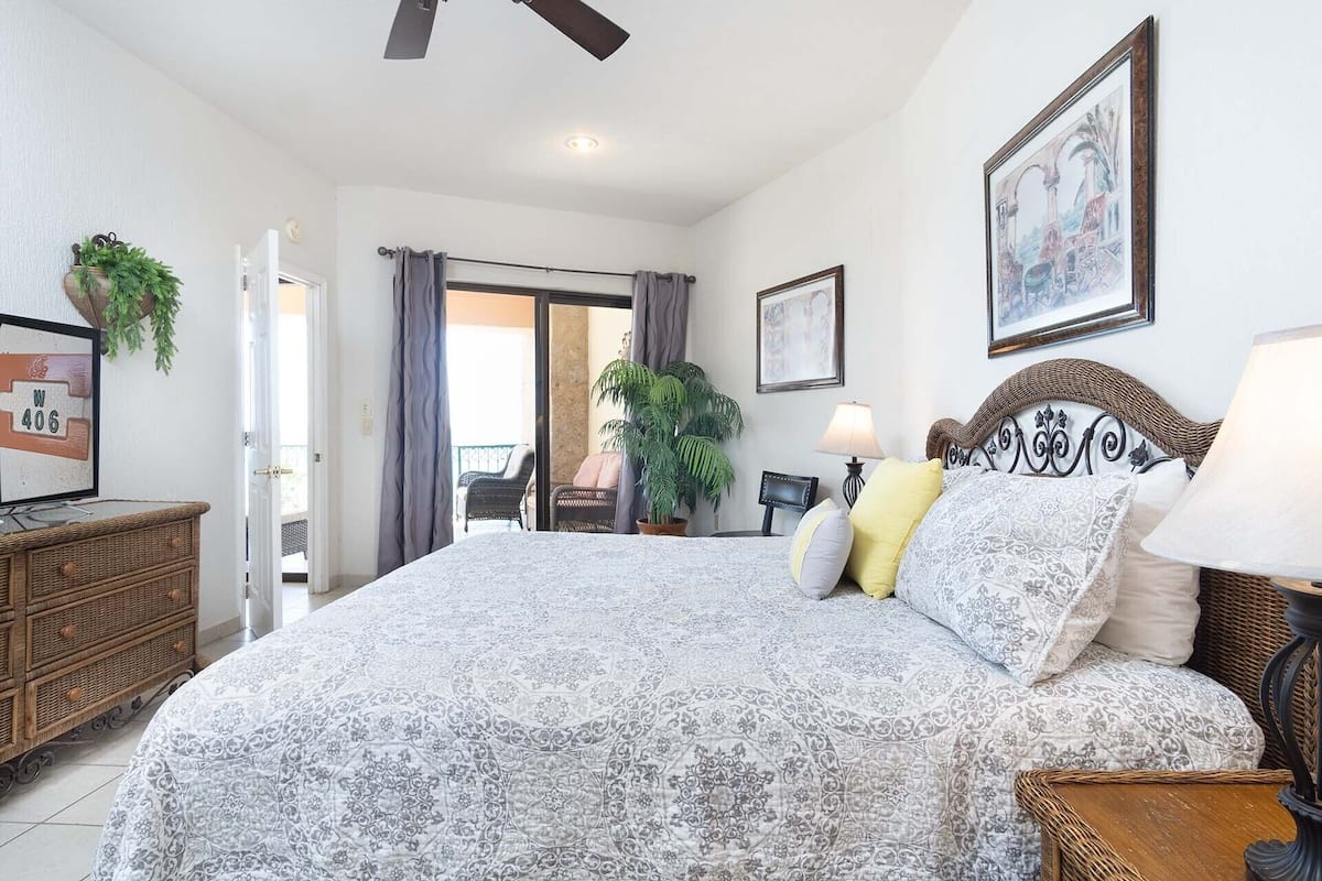 A bedroom with a large bed, a television, a wicker side table, a potted plant, and a balcony with a chair.