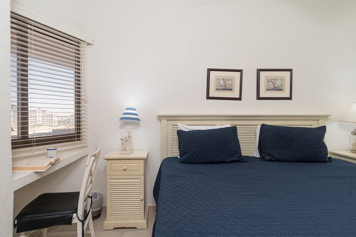 A bedroom with a bed, two bedside tables, a window with blinds, a chair, and two framed pictures on the wall.