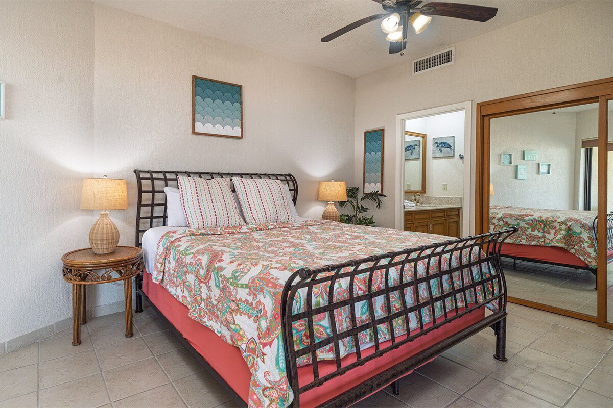 A bedroom with a bed, two bedside tables, a ceiling fan, a large mirror, and a wall with framed artwork.