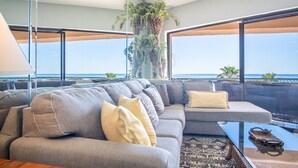 A modern living room with a grey sofa, a glass coffee table, and a view of the ocean.