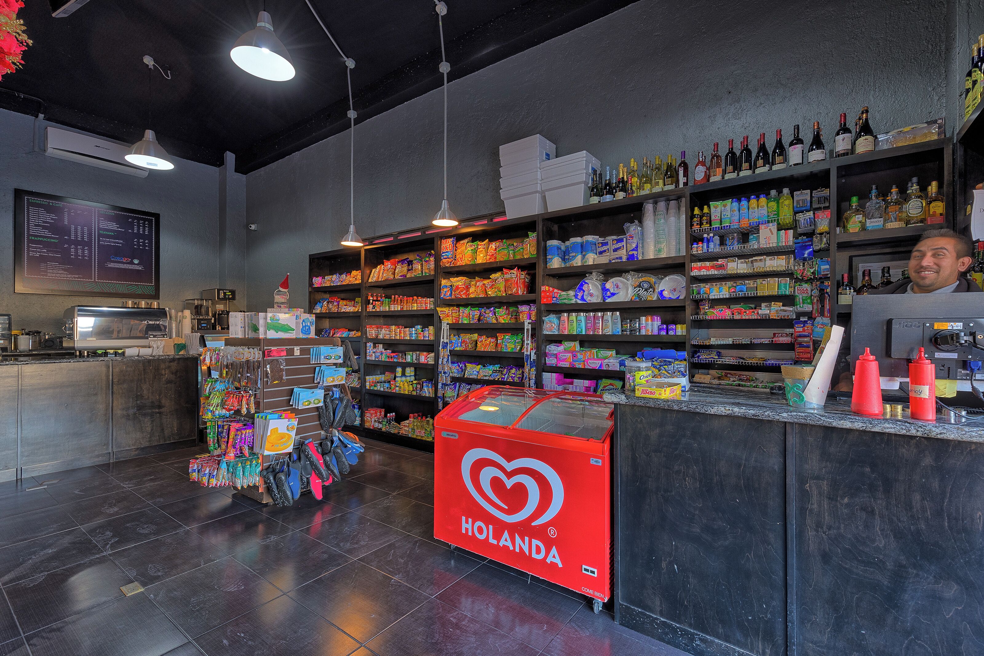 A store interior with shelves stocked with various products, a refrigerated display case, and a digital screen on the wall.