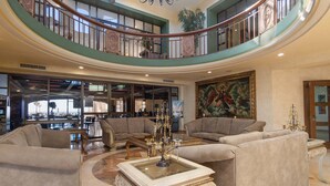 A spacious lobby with a high ceiling, a grand staircase, and a large painting. There are two sofas and a coffee table in the center.