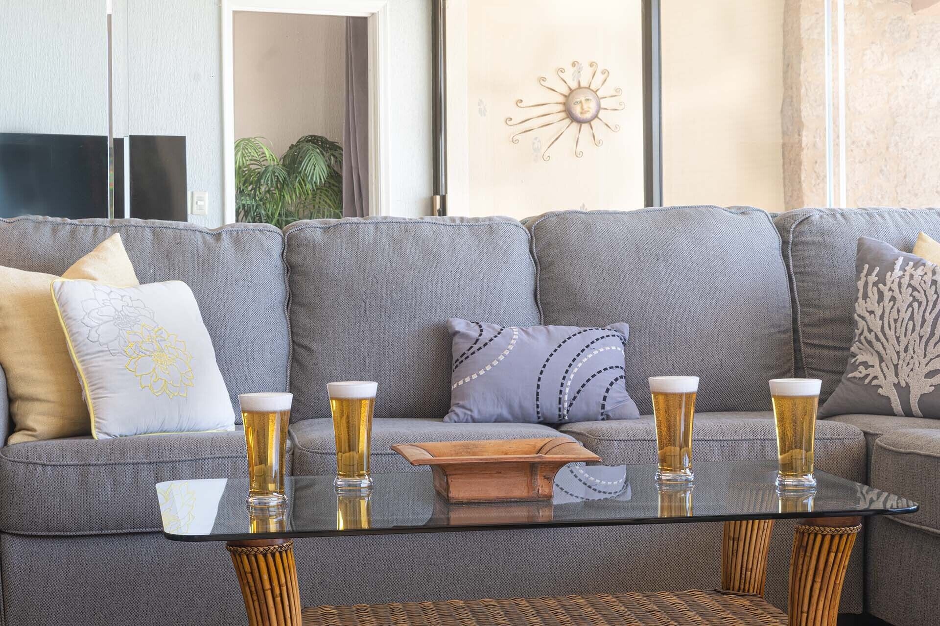 Un salón con un sofá gris, dos almohadones decorativos, una mesa de centro de vidrio y cuatro vasos de cerveza, y una pequeña bandeja de madera.