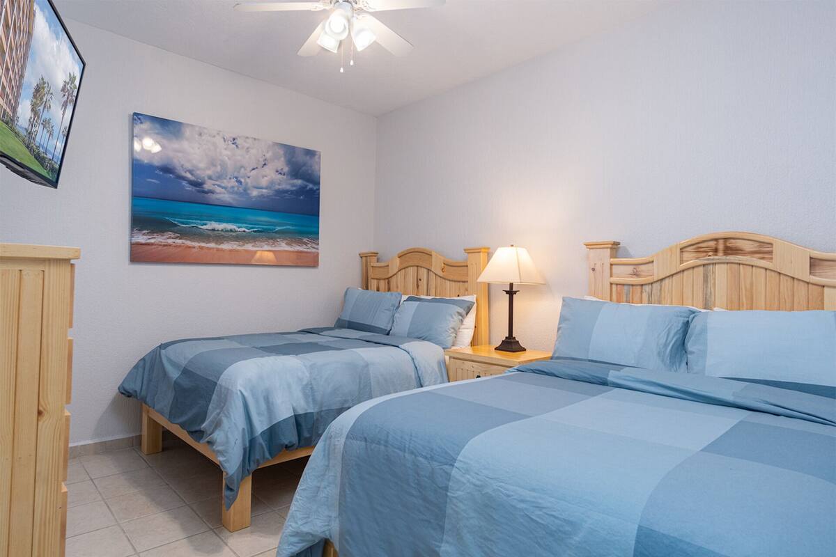 Ein Schlafzimmer mit zwei Betten, einem Nachttisch mit Lampe, einem Holz-Kopfteil, einem Bild mit Strandmotiv und einem Deckenventilator.