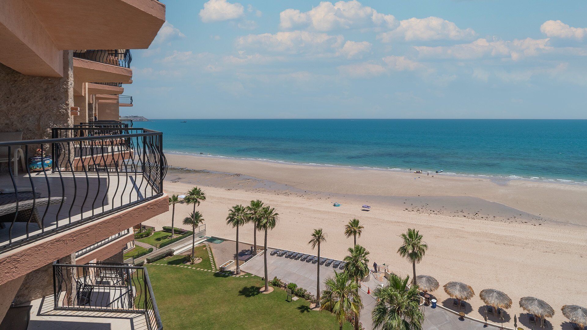 Un hôtel situé en bord de mer, doté d’un balcon avec vue sur l’océan, d’une plage de sable et de palmiers.