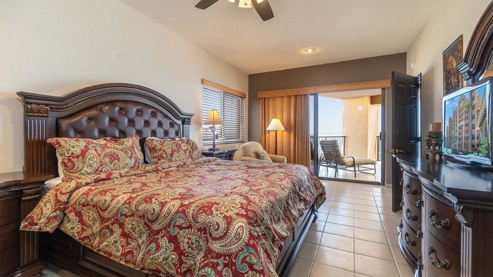 Un'ampia camera da letto con un grande letto, una cassettiera, una televisione e un balcone con una sedia.