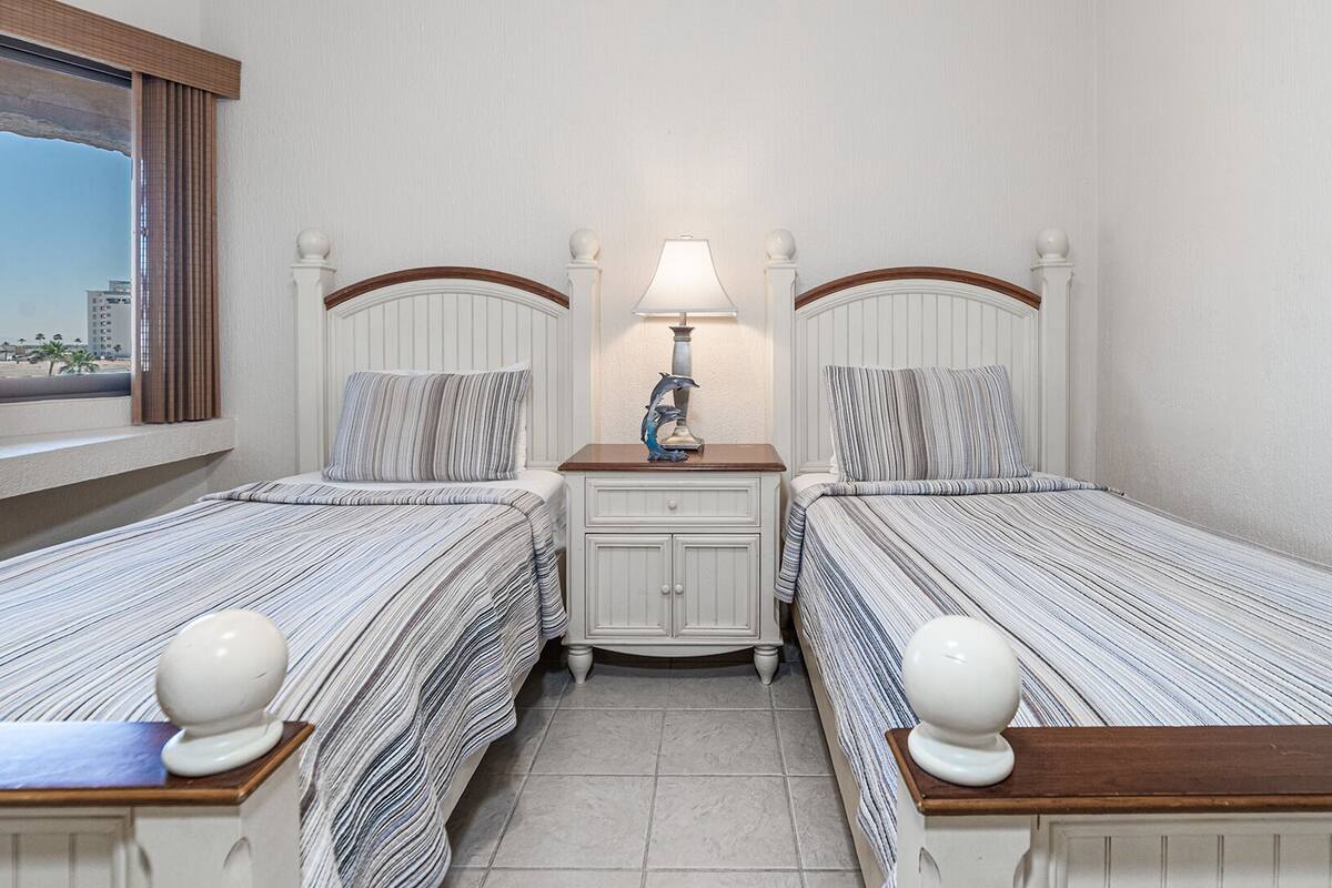 Una habitación de hotel con dos camas individuales, una mesita de noche con lámpara y una ventana con vistas a edificios.