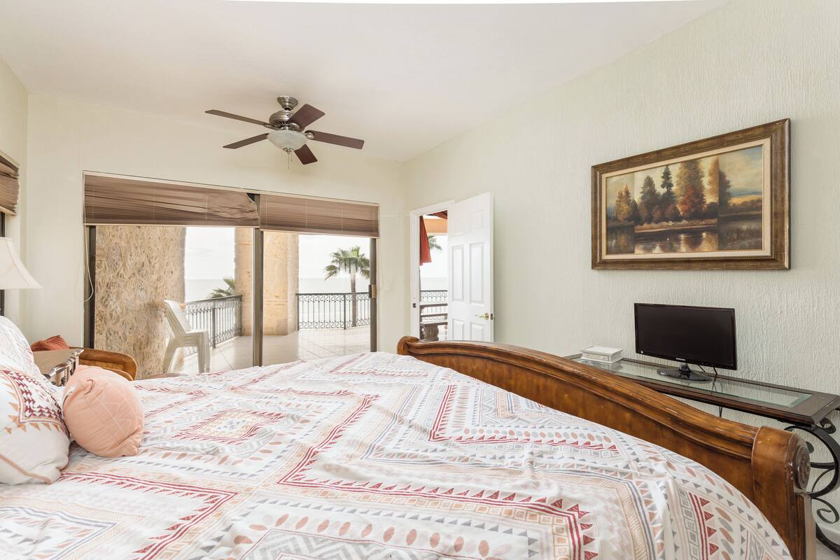 Un dormitorio con cama, ventilador de techo, un cuadro, escritorio con computadora y vistas al exterior.