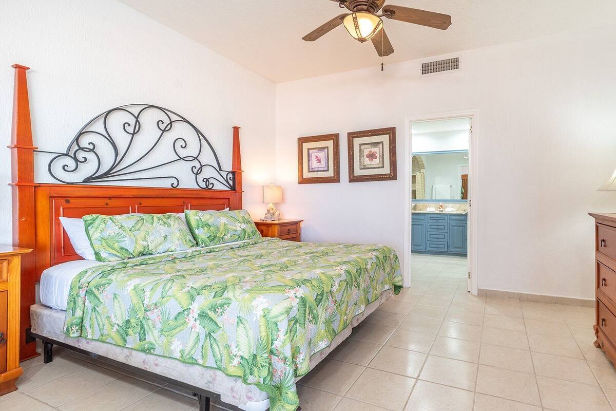 Un dormitorio con cama, cabecera de madera, ventilador de techo, televisor y vistas al baño.