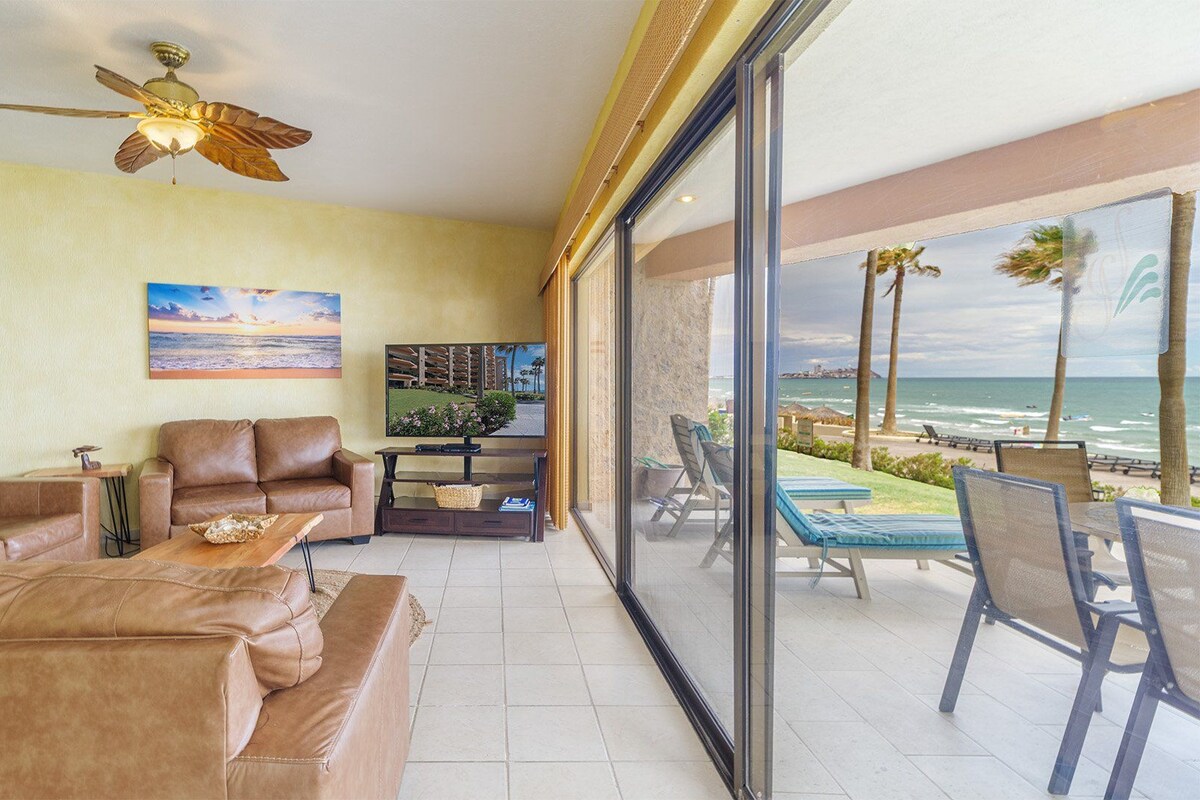 A living room with a large window overlooking a beach, a ceiling fan, and a television on a stand.