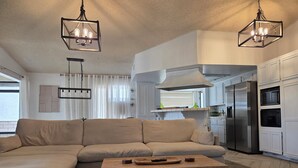 A modern living room with a beige sectional sofa, a wooden coffee table, and a kitchen area with white cabinets and stainless steel appliances. - Minimalist Lake House (Helendale)