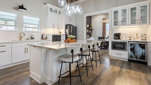 Une cuisine moderne avec des meubles blancs, un grand îlot central doté de tabourets, un four encastré et un luminaire suspendu.