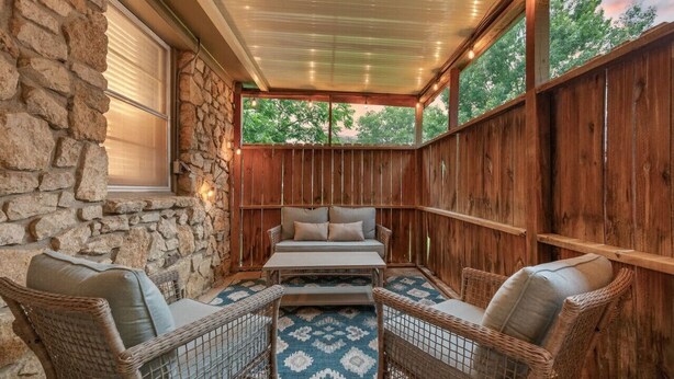 A covered patio with stone walls, wooden paneling, wicker furniture, and a patterned rug. - Rustic Charm Owasso Retreat | 2BR • Big Backyard (Owasso)
